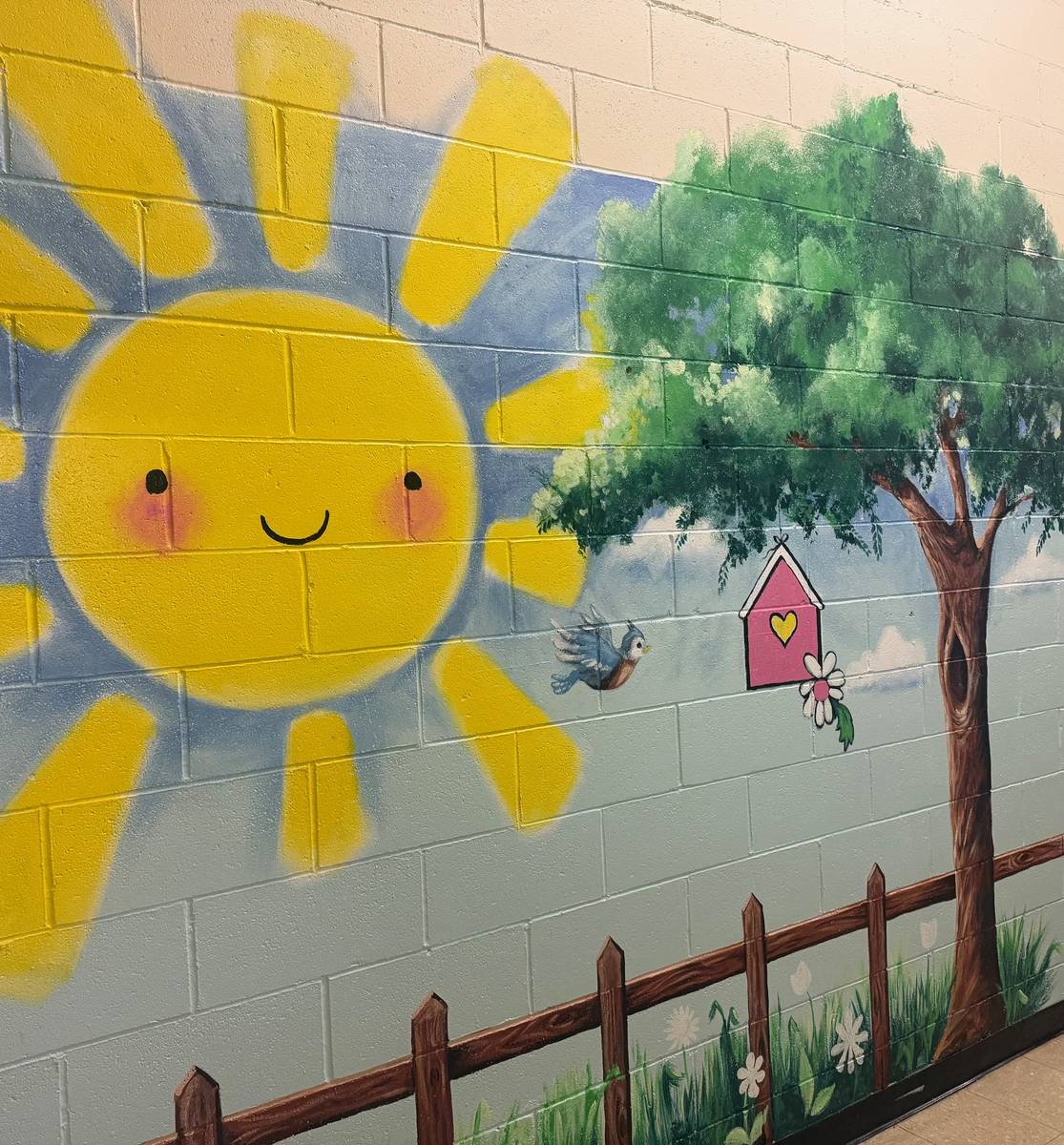 A hand-painted mural of a smiling sun, tree, birdhouse and picket fence in the school hallway